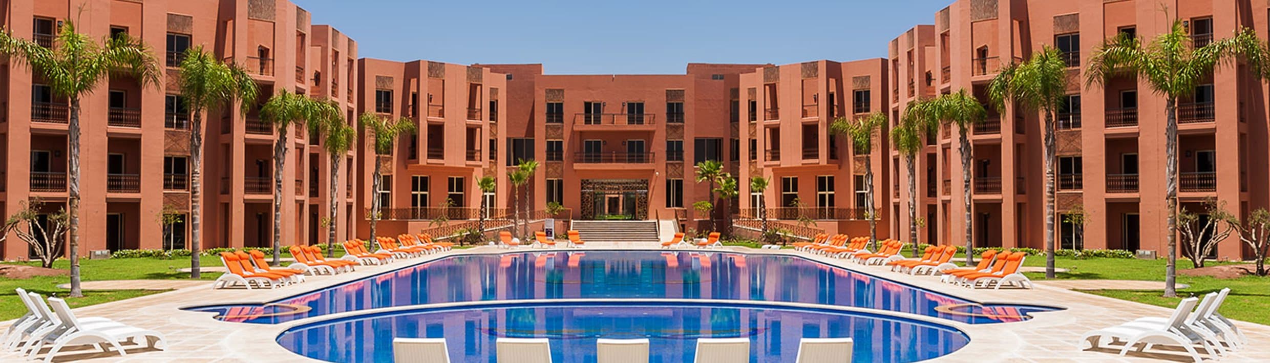 Panoramic view of the pool framed by the hotel's ochre-red buildings